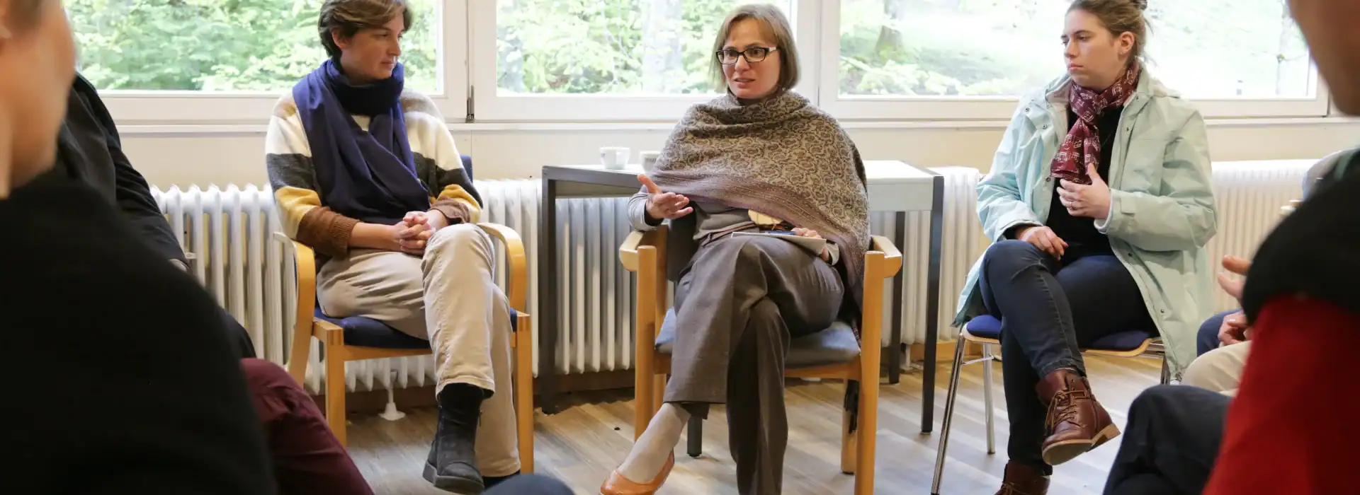 Drei Menschen sitzen vor einer Fensterfront im Kreis, eine Frau spricht und gestikuliert dabei.